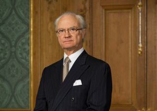 Kungen står framför en stor brun dörr. Han ser allvarlig ut och tittar in i kameran. Han har grått tunt hår, glasögon och kostym med slips.