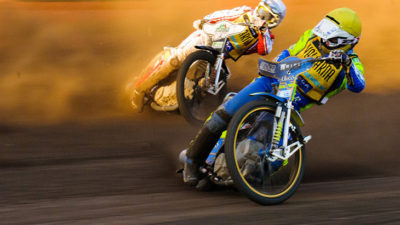 Två speedwaymotorcyklar åker på en grusbana. Båda förarna lutar cyklarna väldigt mycket och det sprutar sand. Båda har mycket skydd och stora hjälmar på sig.