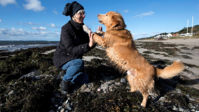 Hunden Nimbus står med tassarna mot sin matte Mariannes händer. Nimbus är ljusbrun med medellång päls. Marianne har blåa jeans och en svart sjal knuten runt huvudet. De står på en strand full med alger.