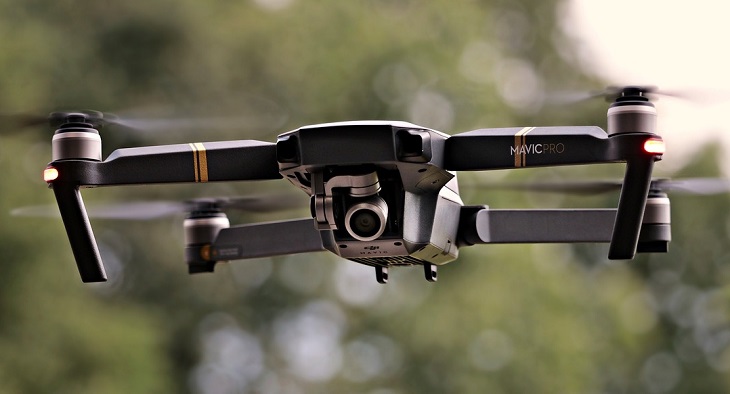 En drönare flyger i luften. Den är grå med fyra pinnar som sticker ut åt varsitt håll. Längst fram syns en kameralins.