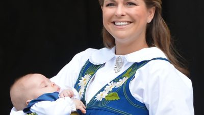 Prinsessan Madeleine har svensk folkdräkt på sig som är gul och blå. Hon ler stort och håller en bebis i sina armar.