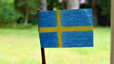 En svensk flagga är ritad med kritor och utklippt. Flaggan sitter på en träpinne. I bakgrundens är det en solig gräsmatta.