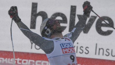 Aksel har skidklädsel, hjälm och skidglasögon på sig. I händerna håller han sina stavar som han sträcker upp i luften som en segergest.